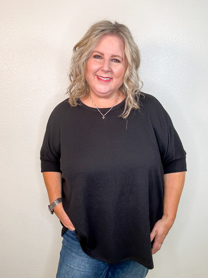 A woman smiling, wearing a black dolman sleeve top with a rounded neckline and short drop shoulder sleeves, paired with jeans.