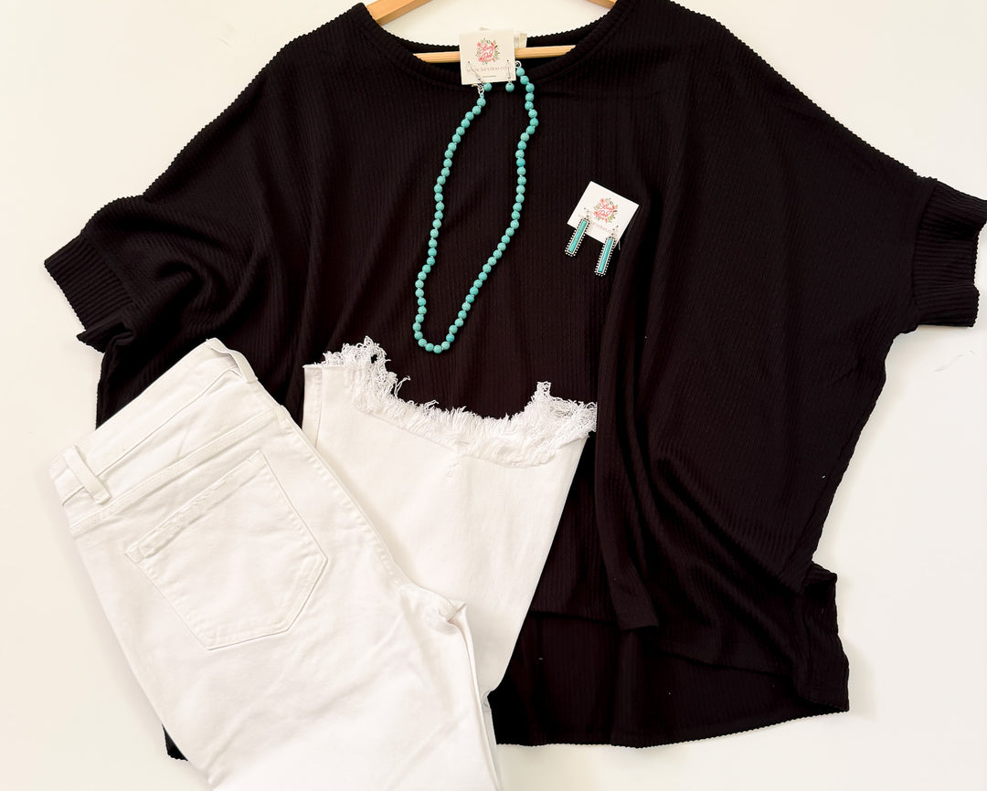 A black, dolman-sleeved, waffle knit top laid out flat with white denim shorts, a turquoise necklace, and a tag visible in the image.