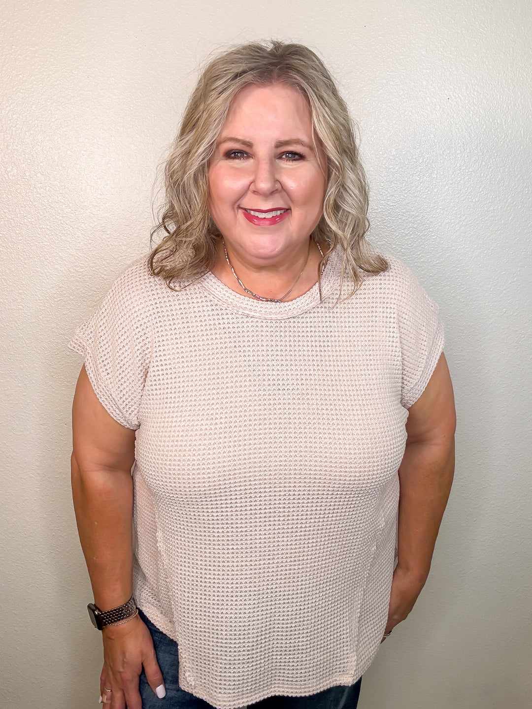 A smiling woman wearing a beige, short-sleeved, waffle knit top with a round neckline and hi-low hemline.