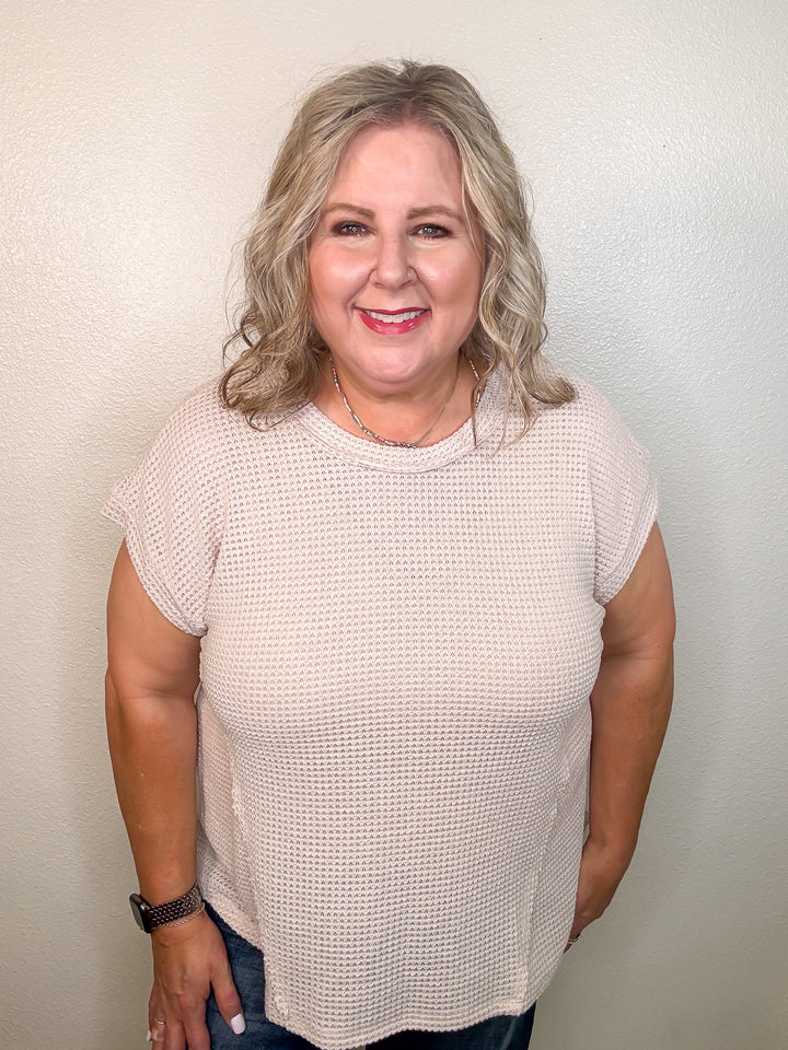 A smiling woman wearing a beige, short-sleeved, waffle knit top with a round neckline and hi-low hemline.