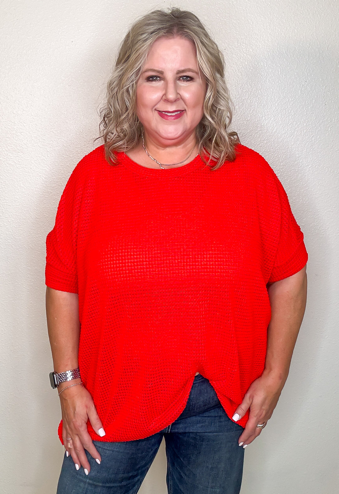 A person wearing a red, oversized, short-sleeved crochet top with a round neckline, paired with dark jeans.