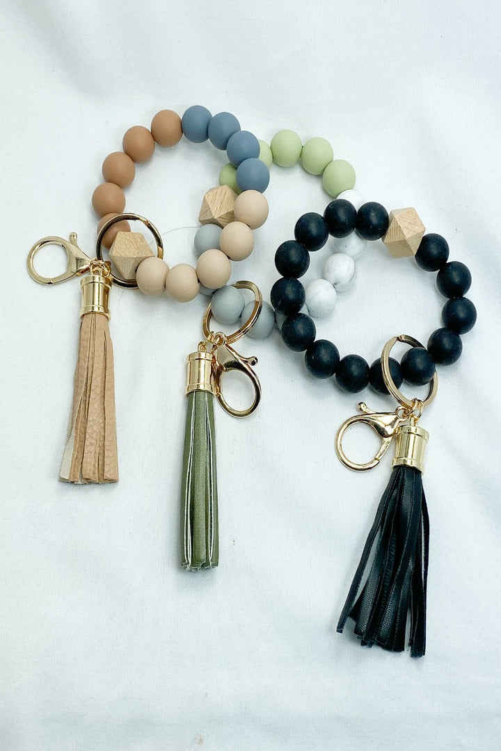 A collection of round keychains with tassels in various colors including beige, black, blue, and green, arranged on a white surface.