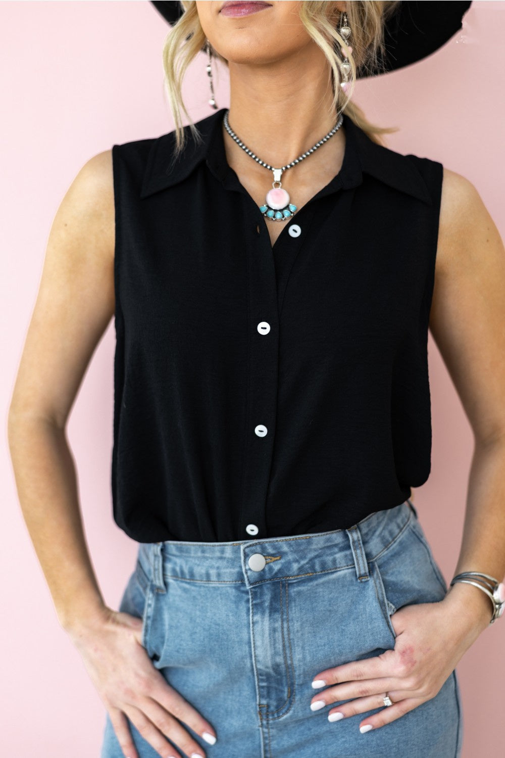 A person wearing a black sleeveless button-down blouse with a collar, paired with blue denim shorts and accessorized with a necklace and watch.