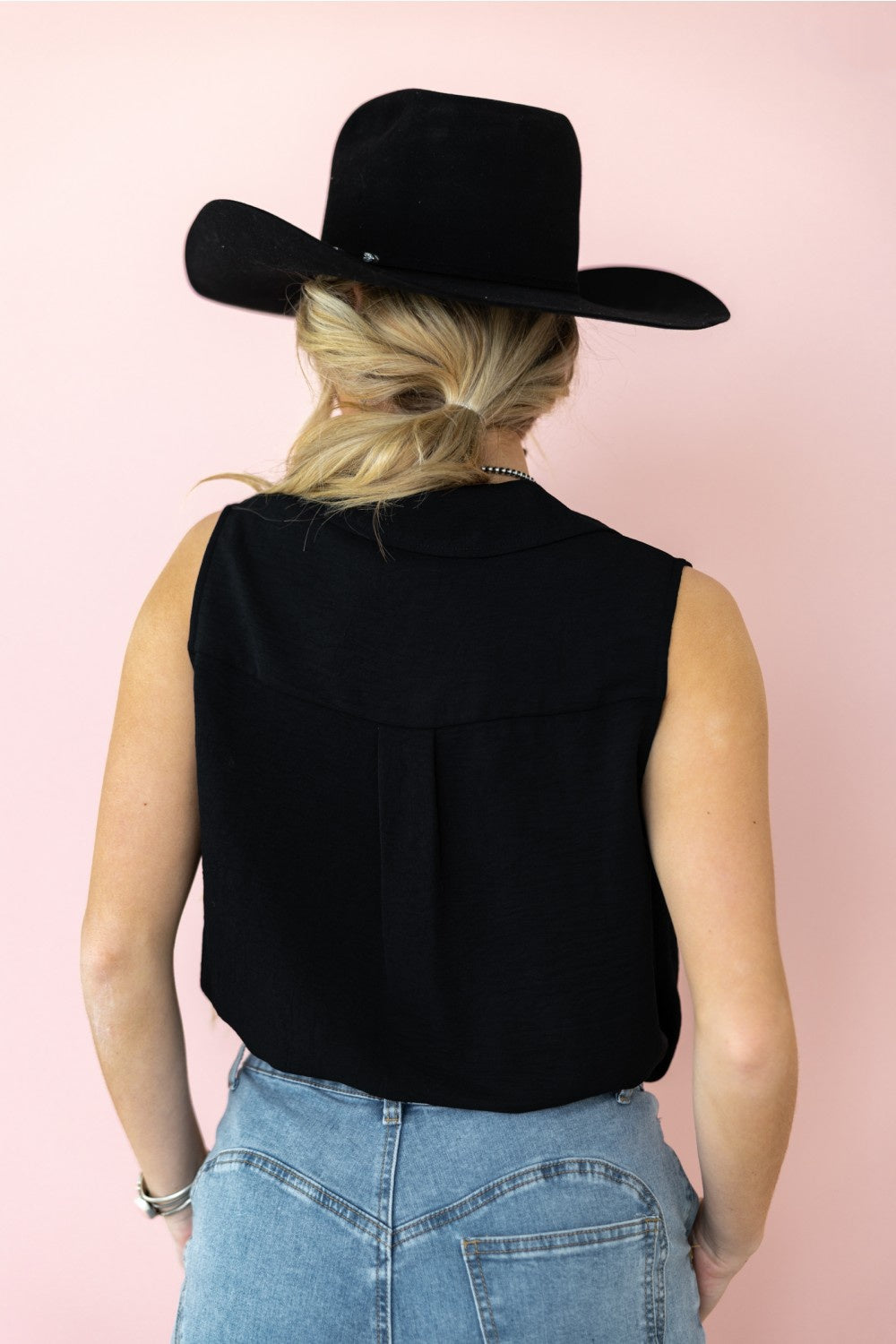 Person wearing a black sleeveless top and wide-brimmed black hat on a pink background