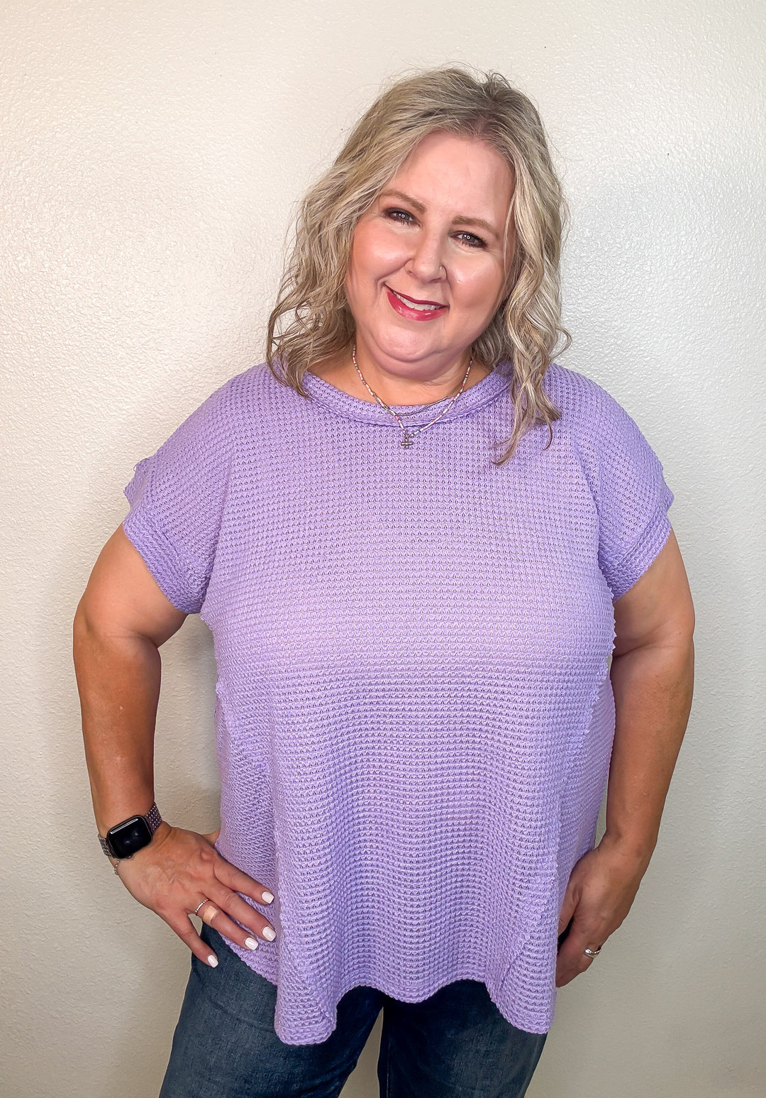 A person wearing an orchid purple, short-sleeved, waffle knit top with a round neckline and hi-low hemline, paired with jeans.