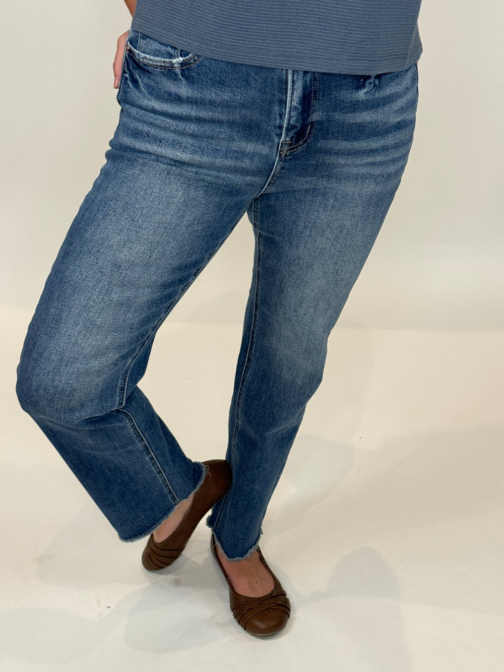 A person standing against a white background, wearing The Alex Jean which is a high-rise straight-leg blue denim jean and brown shoes.