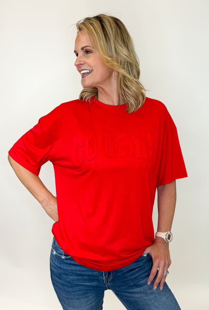 A person wearing a red 'GO TEAM' t-shirt with embossed text on the front, paired with blue jeans.