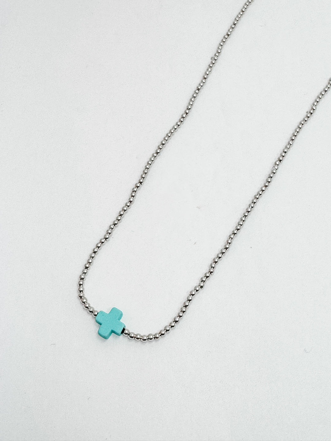 A silver necklace with a turquoise cross accent, featuring petite matte silver beads and a simple turquoise square cross.